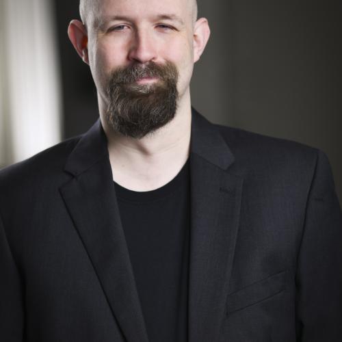 A bald caucasian man with a dark goatee/beard. He is wearing a dark shirt and suit jacket